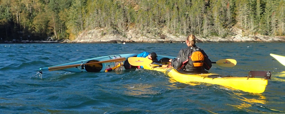 sea kayaking rescue practice: one sea kayaker offers the bow of her kayak to a capsized paddler, so the upside down paddler can roll up without having to wet exit their boat.